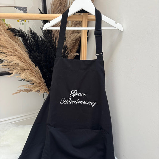 Black apron with 'Grace Hairdressing' text hanging on a white wall.