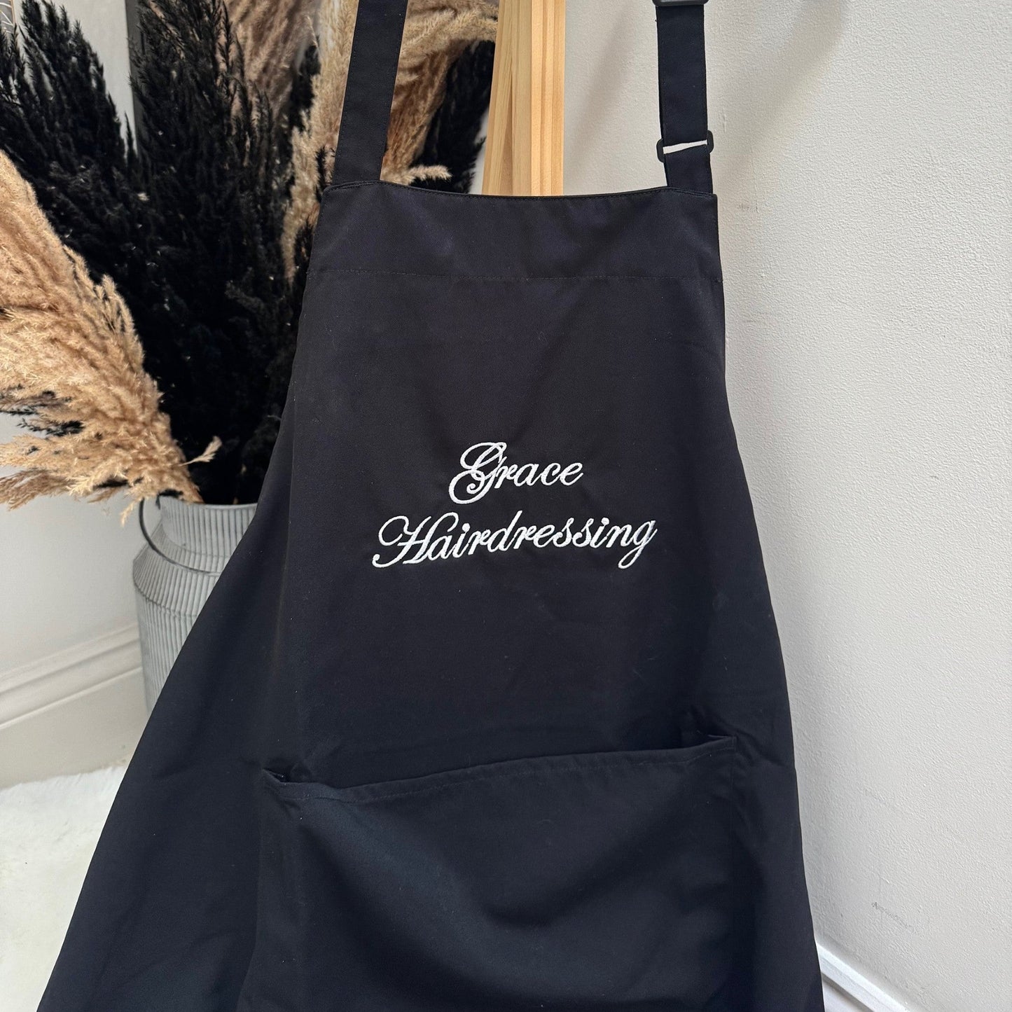 Black apron with 'Grace Hairdressing' text hanging on a white hanger against a neutral background.