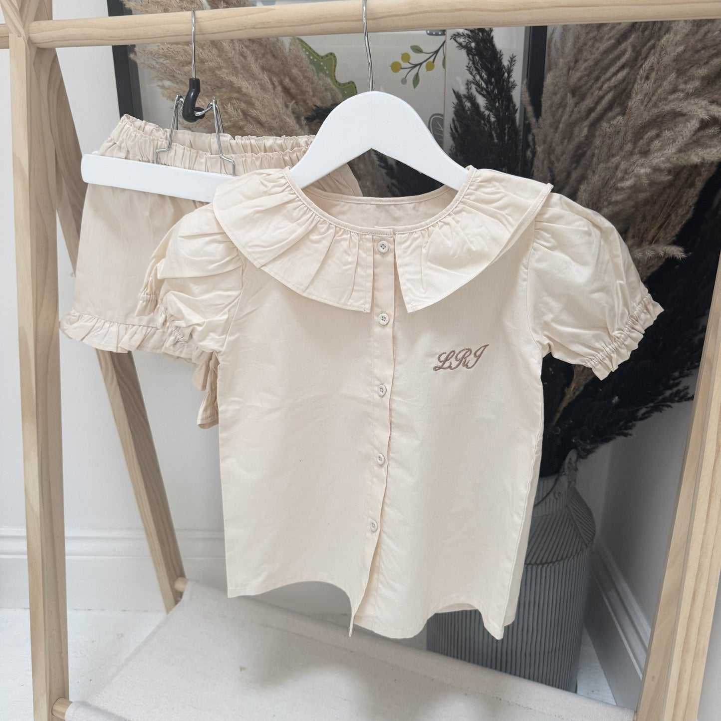 Beige blouse with personalised initials with ruffled collar hanging on a wooden rack with dried plants in the background.