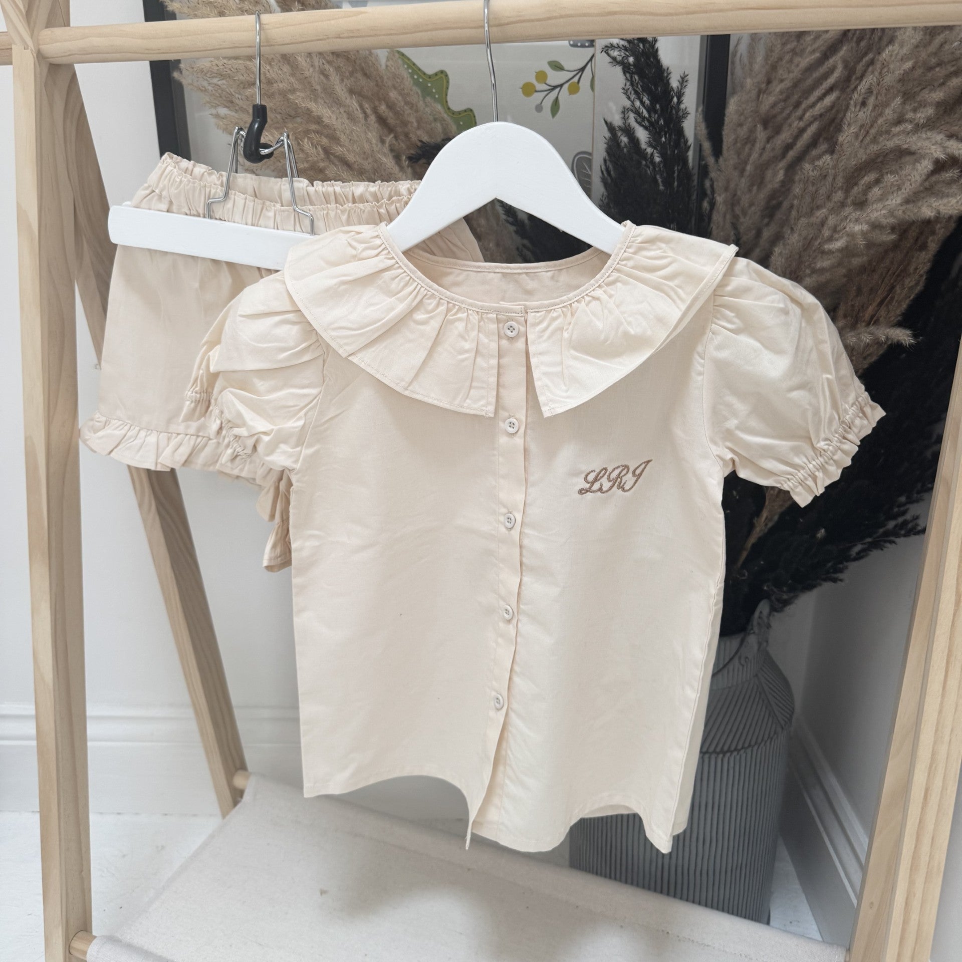 Beige blouse with personalised initials with ruffled collar hanging on a wooden rack with dried plants in the background.