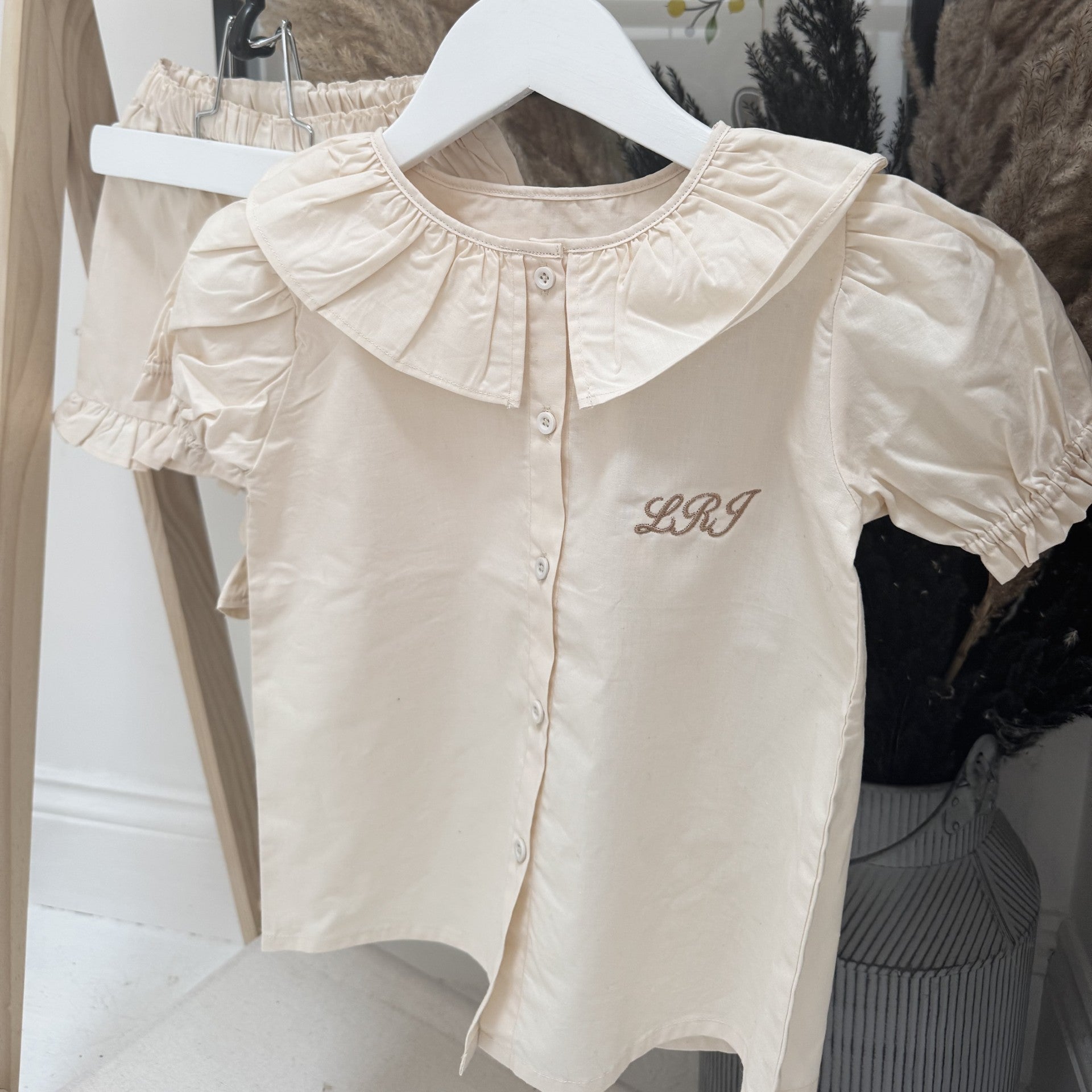 Beige blouse with ruffled collar and 'L.R.J.' embroidery on a hanger.