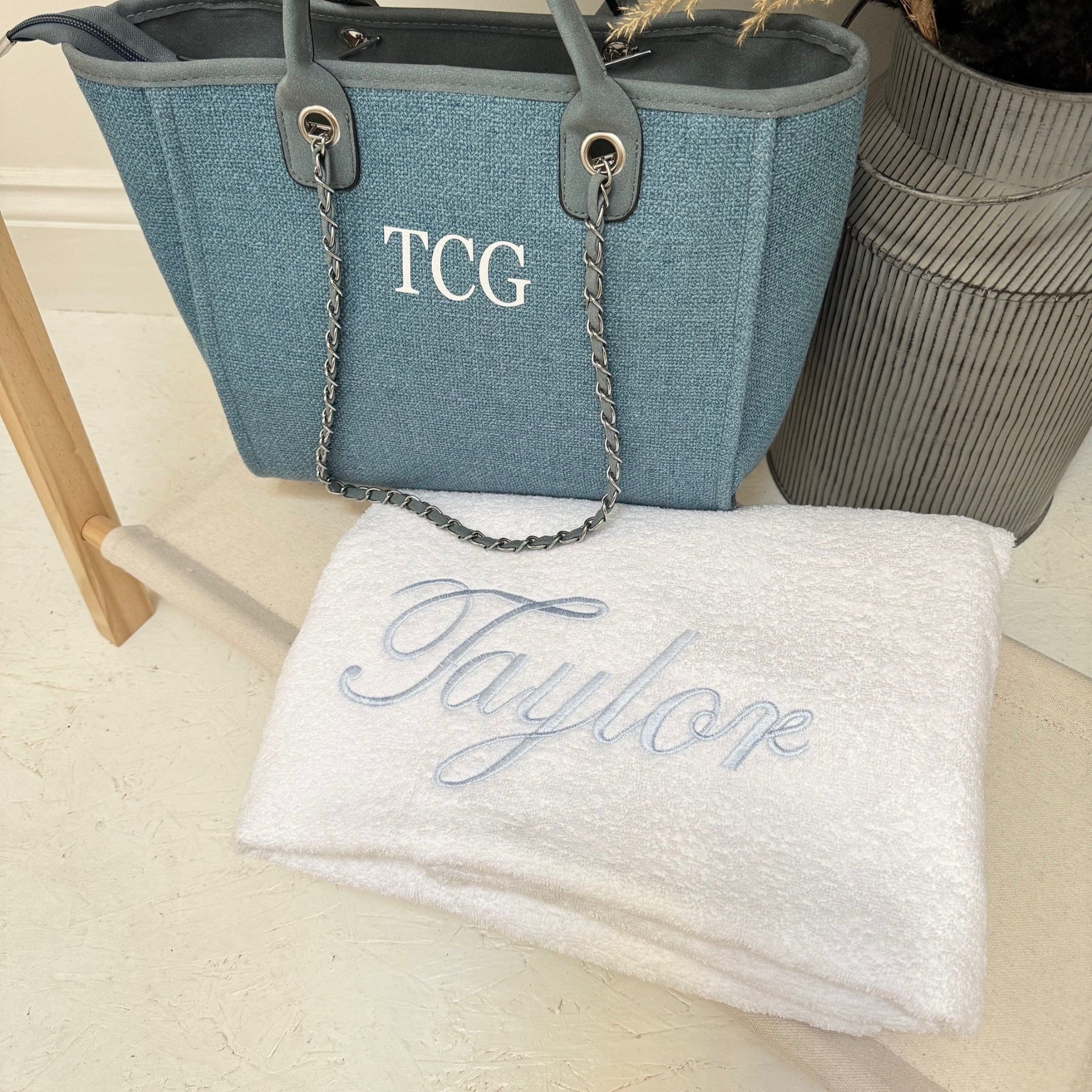 Blue handbag with 'TCG' monogram and white towel with 'Taylor' embroidery on a light wooden surface.