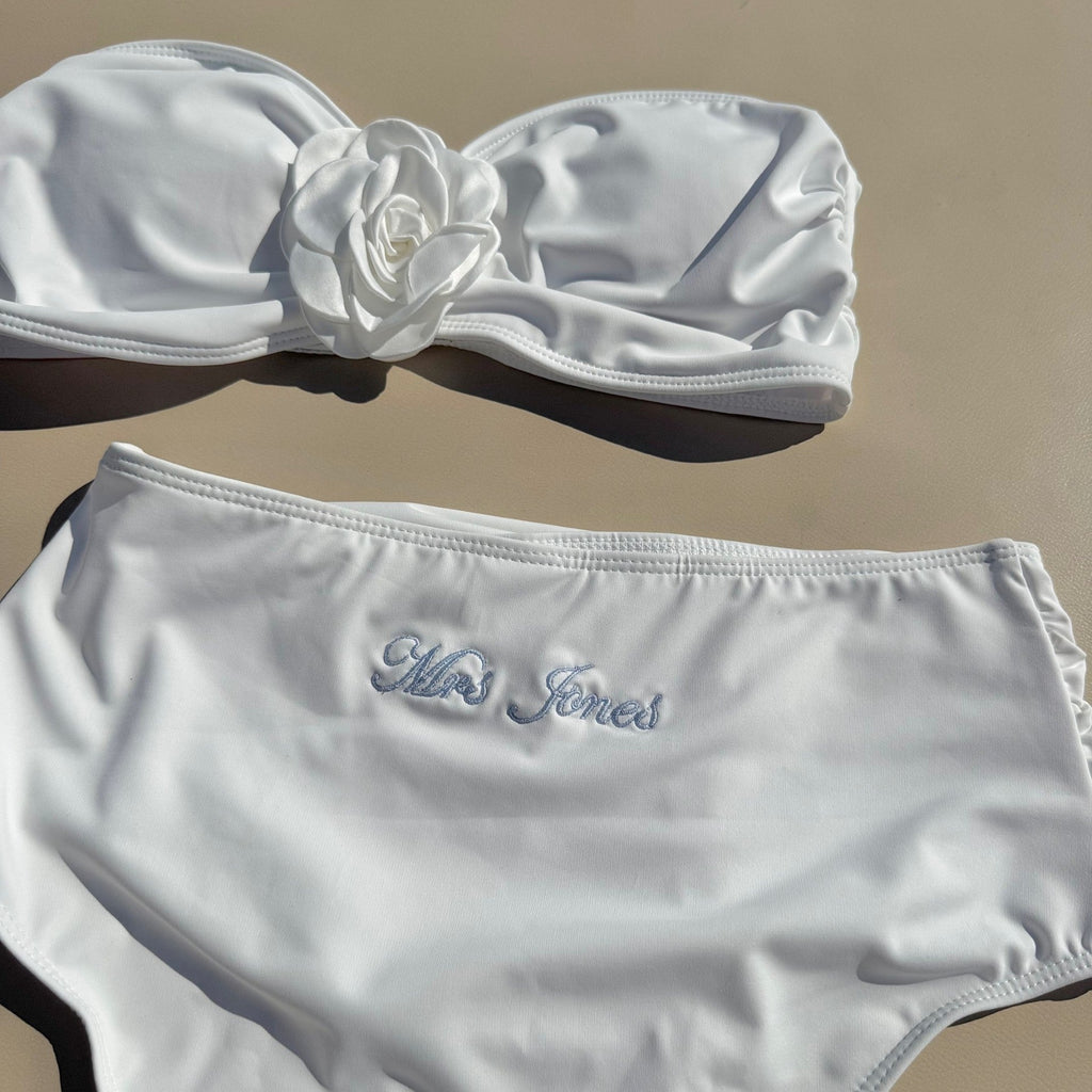White bikini set with floral design on top and bridal name on bottom, against a beige background.