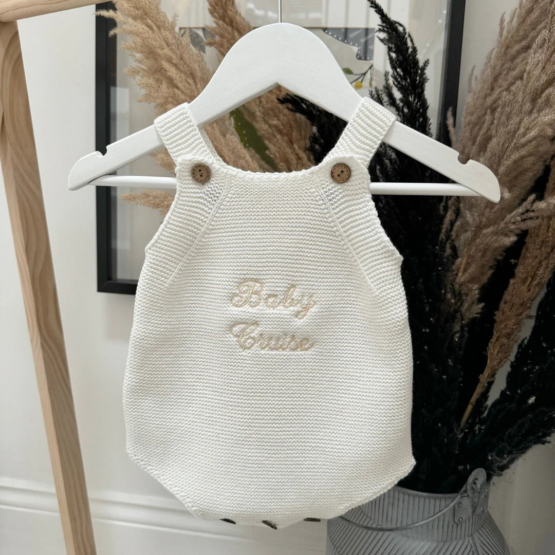 White knitted baby romper with 'Baby Cruise' text on a hanger against a neutral background.