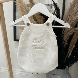 White knitted baby romper with 'Baby Cruise' text on a hanger against a neutral background.