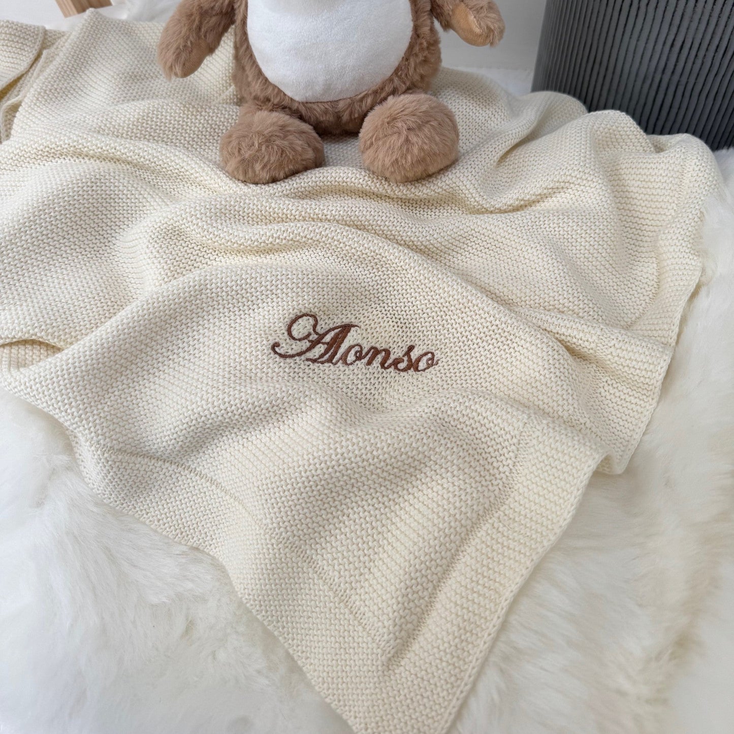 Teddy bear sitting on a white blanket with 'Alonso' embroidered on it, in a room setting.