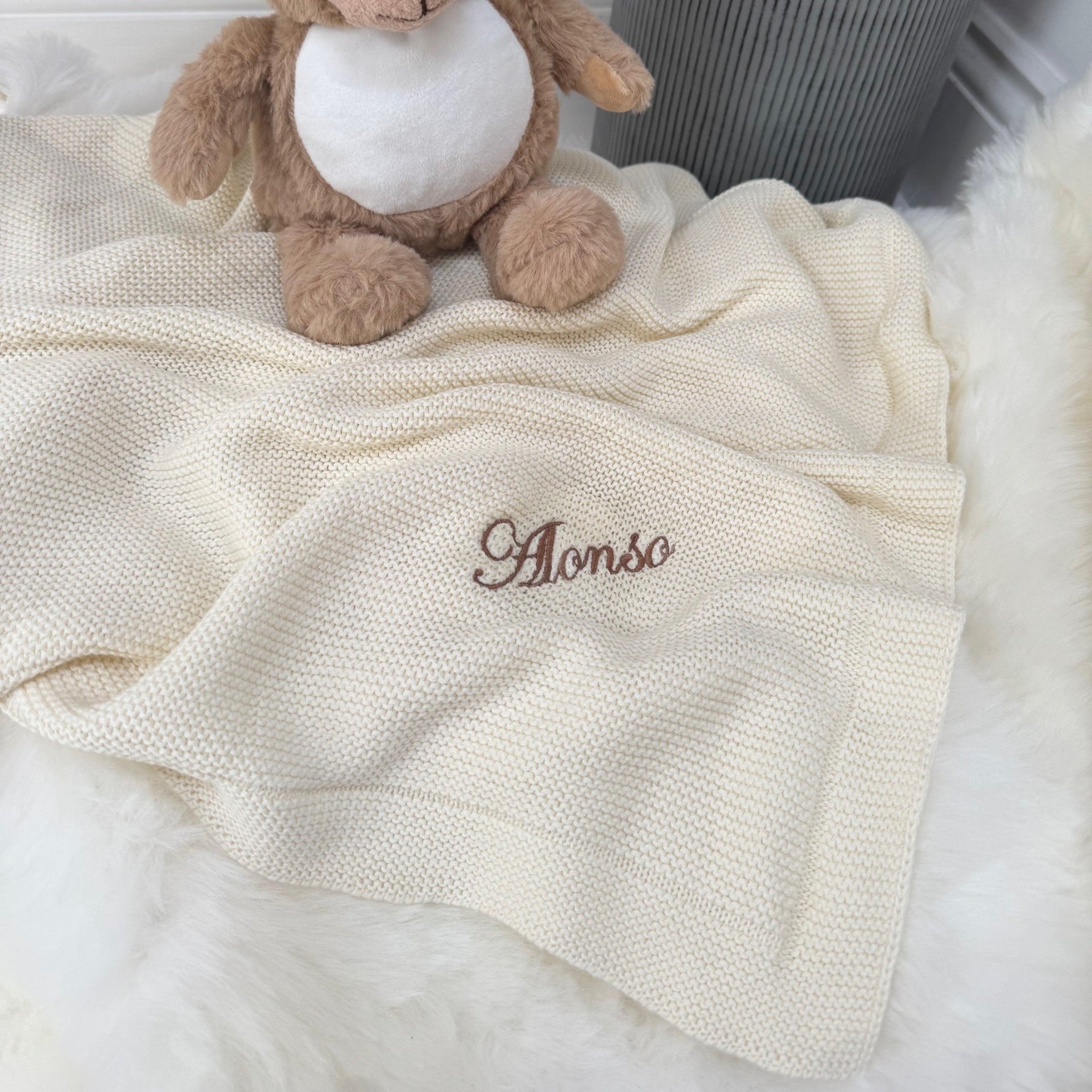 Teddy bear on a white blanket with 'Alonso' embroidered on it, against a neutral background.