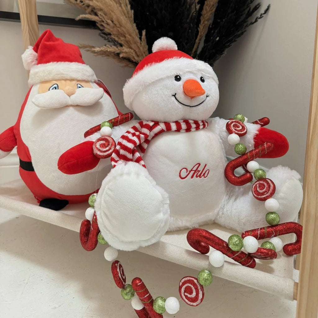Two Christmas-themed plush toys, one resembling Santa Claus and the other a snowman, sitting on a shelf.