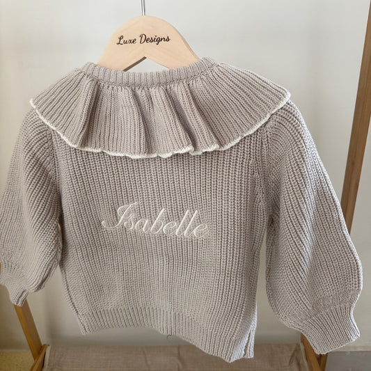 Beige knit sweater with ruffled collar and embroidered name on a wooden hanger against a white wall.