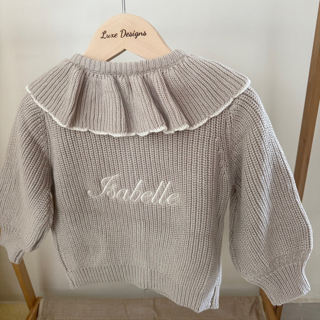 Beige knit sweater with ruffled collar and embroidered name on a wooden hanger against a white wall.