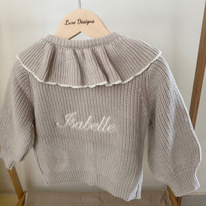 Beige knit sweater with ruffled collar and embroidered name on a wooden hanger against a white wall.