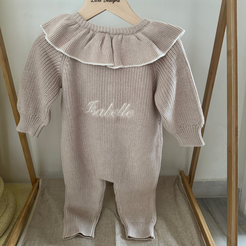 Baby outfit with ruffled collar and embroidered name on a wooden hanger against a white wall.