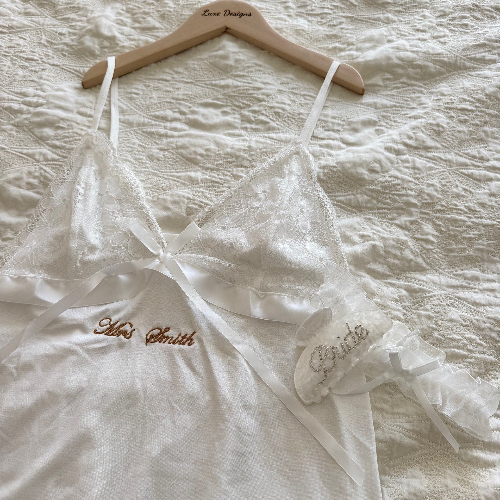 White lace wedding dress with 'Mrs. Smith Bride' embroidery on a wooden hanger over a textured white fabric background.