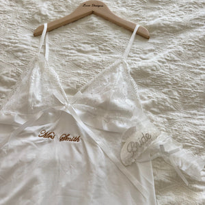 White lace wedding dress with 'Mrs. Smith Bride' embroidery on a wooden hanger over a textured white fabric background.