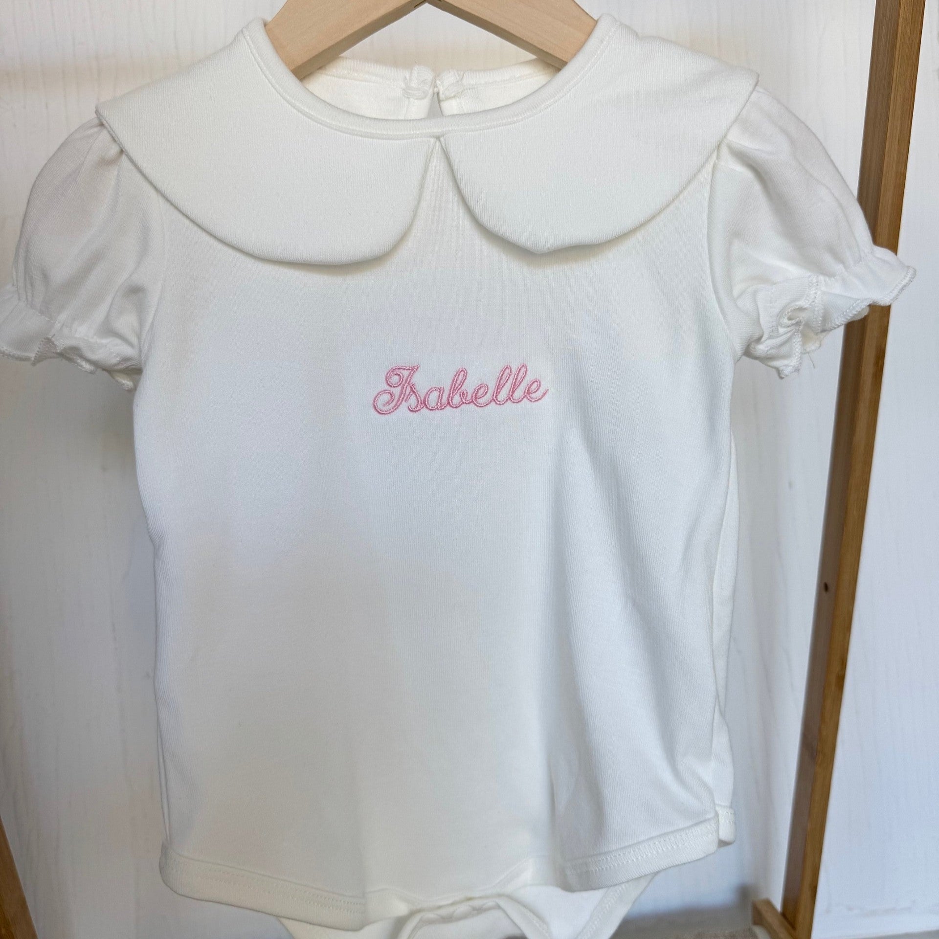 White baby romper with pink embroidery on a wooden hanger against a white background