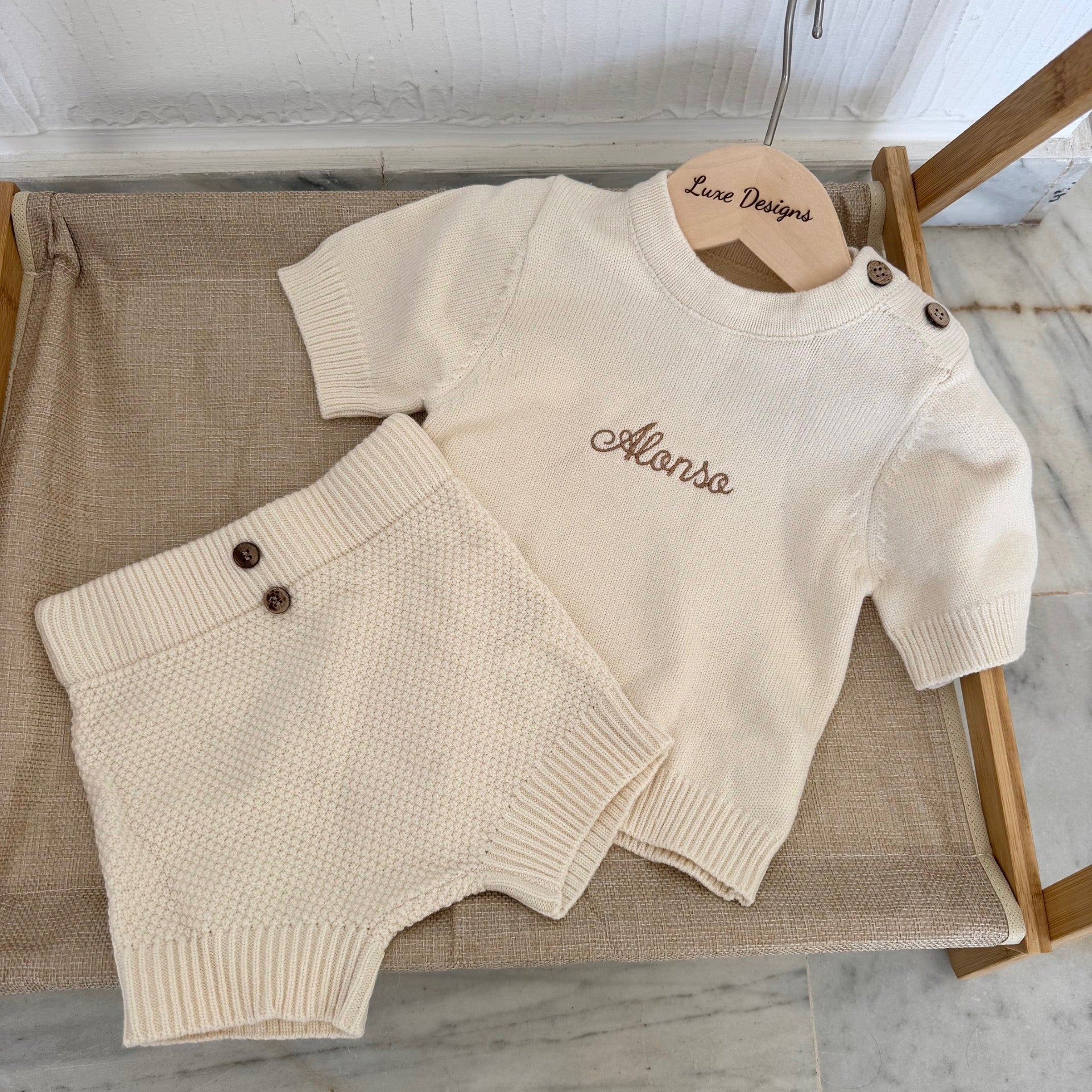 Beige sweater and shorts set on a wooden hanger with 'Alonso' embroidered on it, placed on a textured surface.