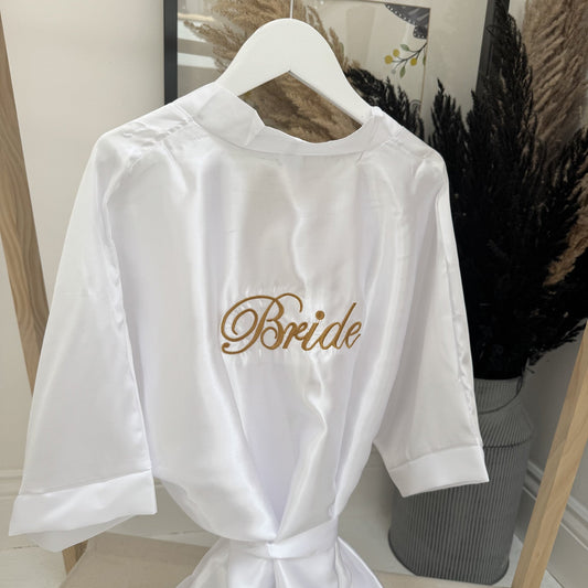 White satin robe with 'Bride' embroidery on a hanger against a neutral background.