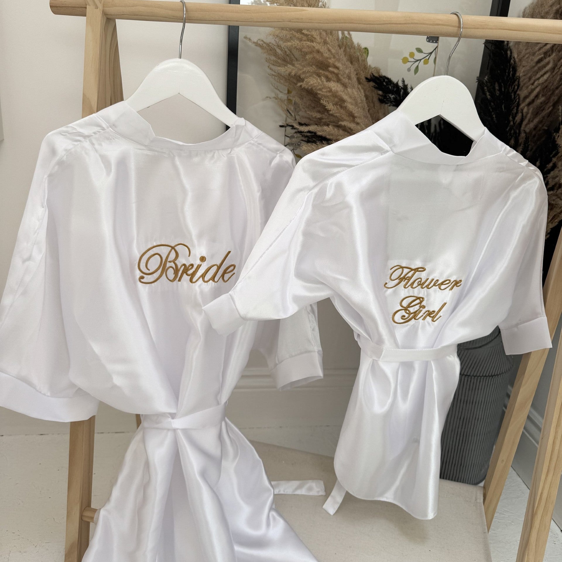 White robes with 'Bride' and 'Flower Girl' text on hangers against a neutral background.