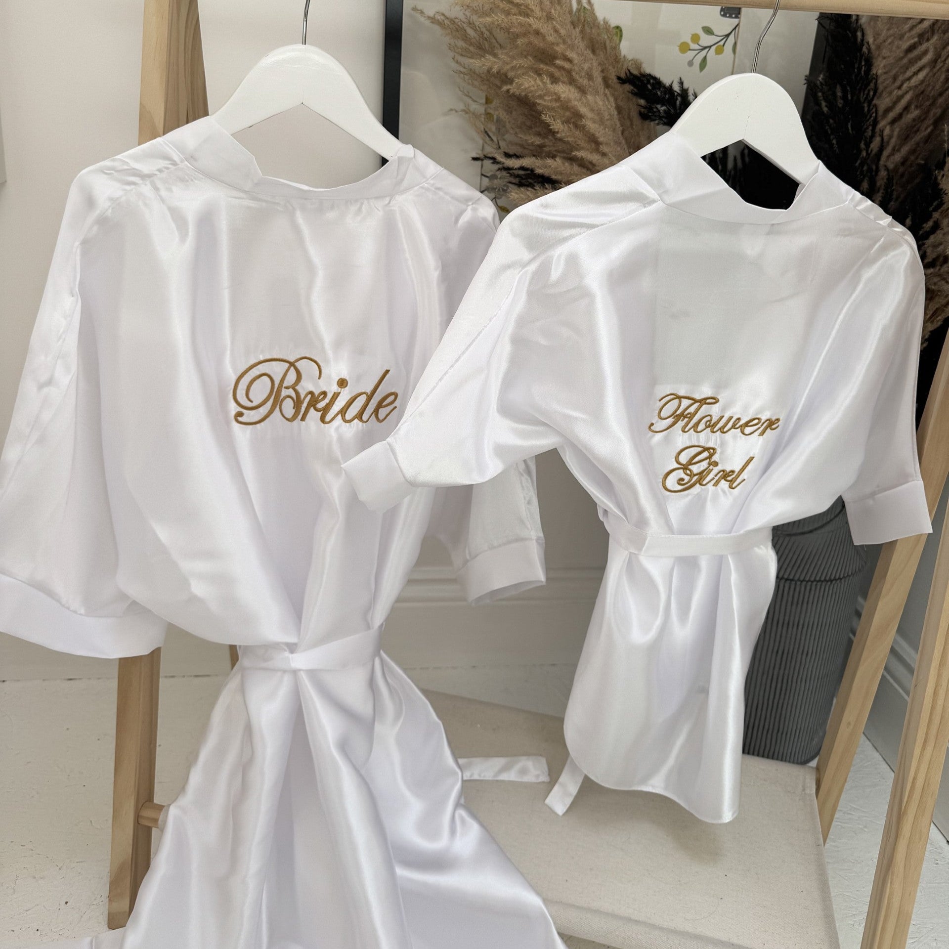 White robes with 'Bride' and 'Flower Girl' text on hangers against a neutral background.