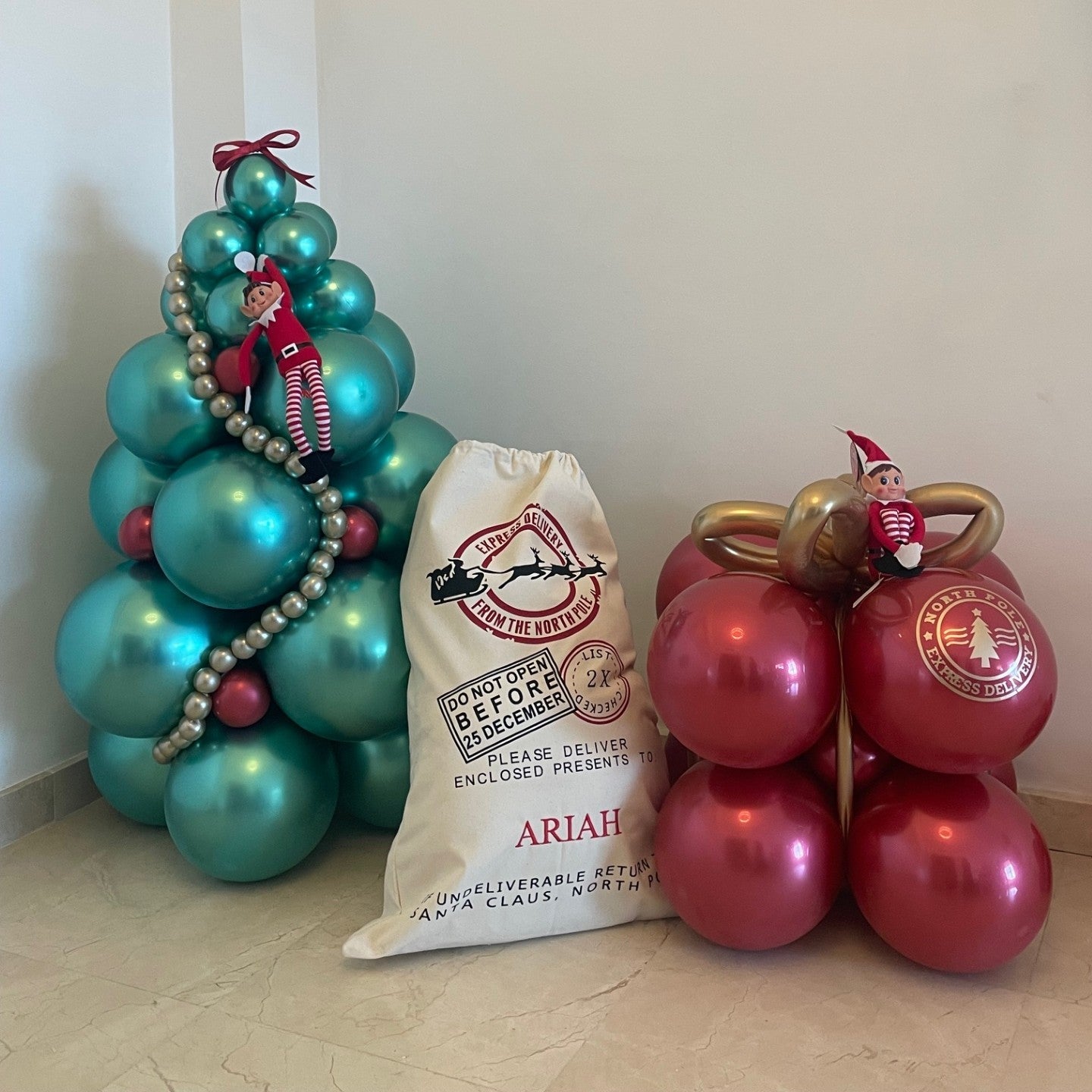 Turquoise balloon tree with red ornaments and a white bag labeled 'Ariah' on a light wooden floor.