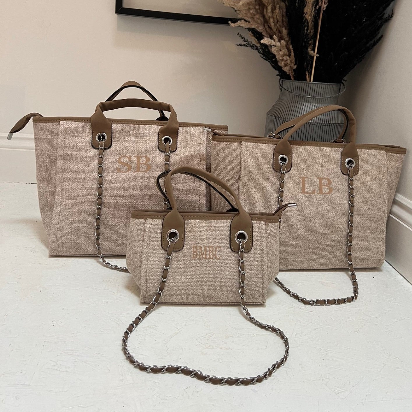 Three brown personalized tote bags, one being a mini one, with shoulder straps and a chain detail, displayed in a home environment.