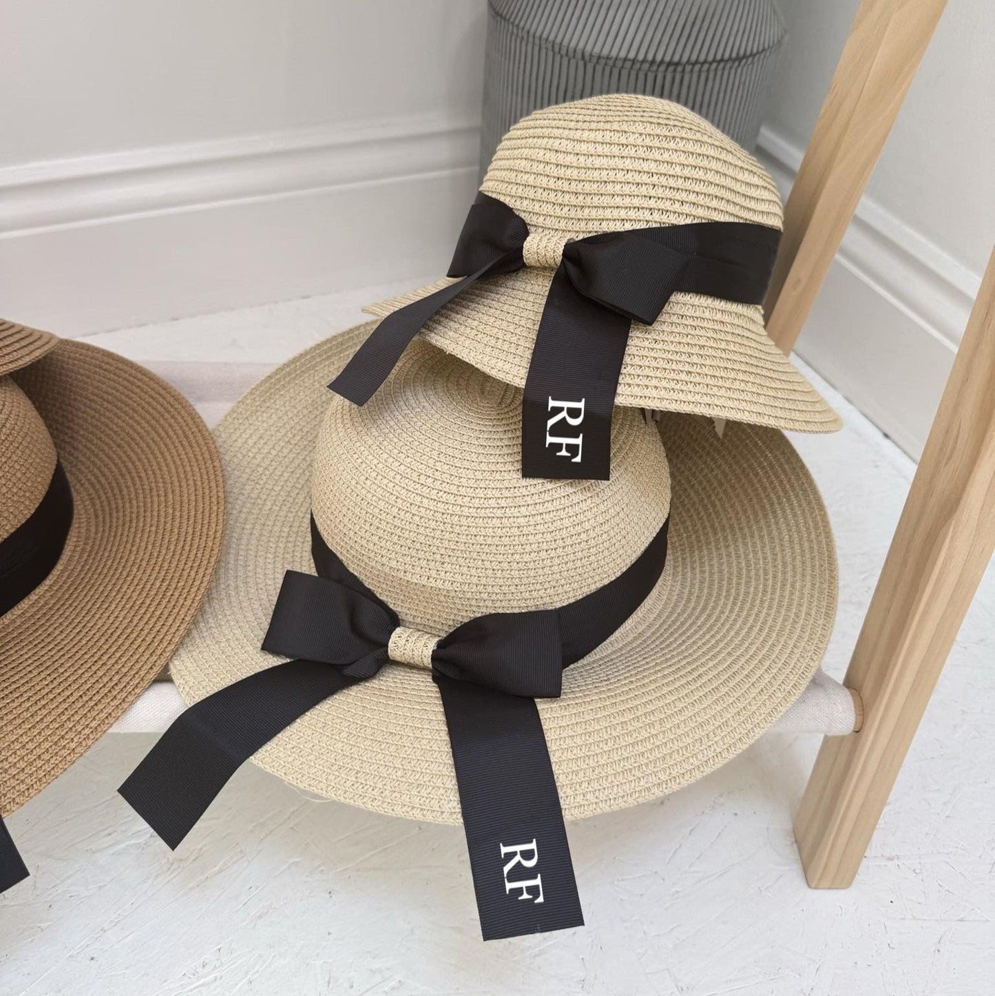 Two straw hats with black ribbons on a wooden stand against a white wall. Both with personalisation