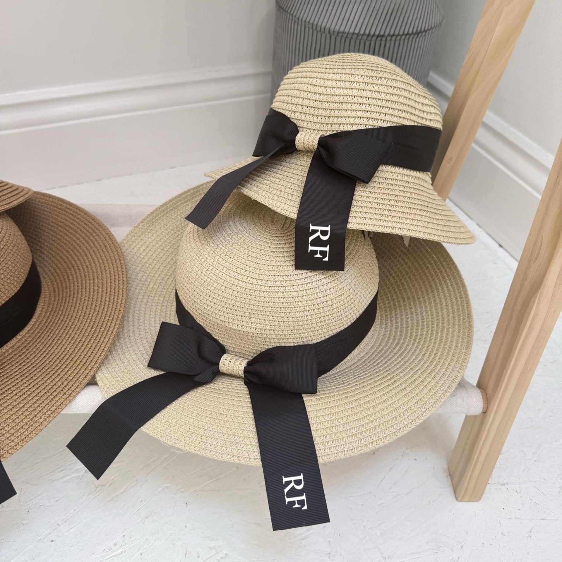 Two straw hats with black ribbons on a wooden stand against a white wall. Both with personalisation
