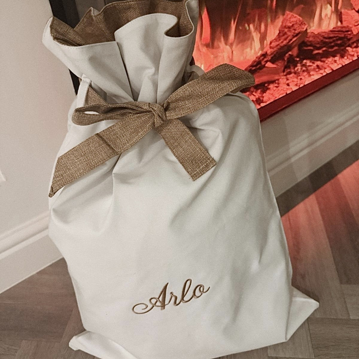 White gift bag with a brown ribbon and 'Arlo' embroidery in front of a fireplace.