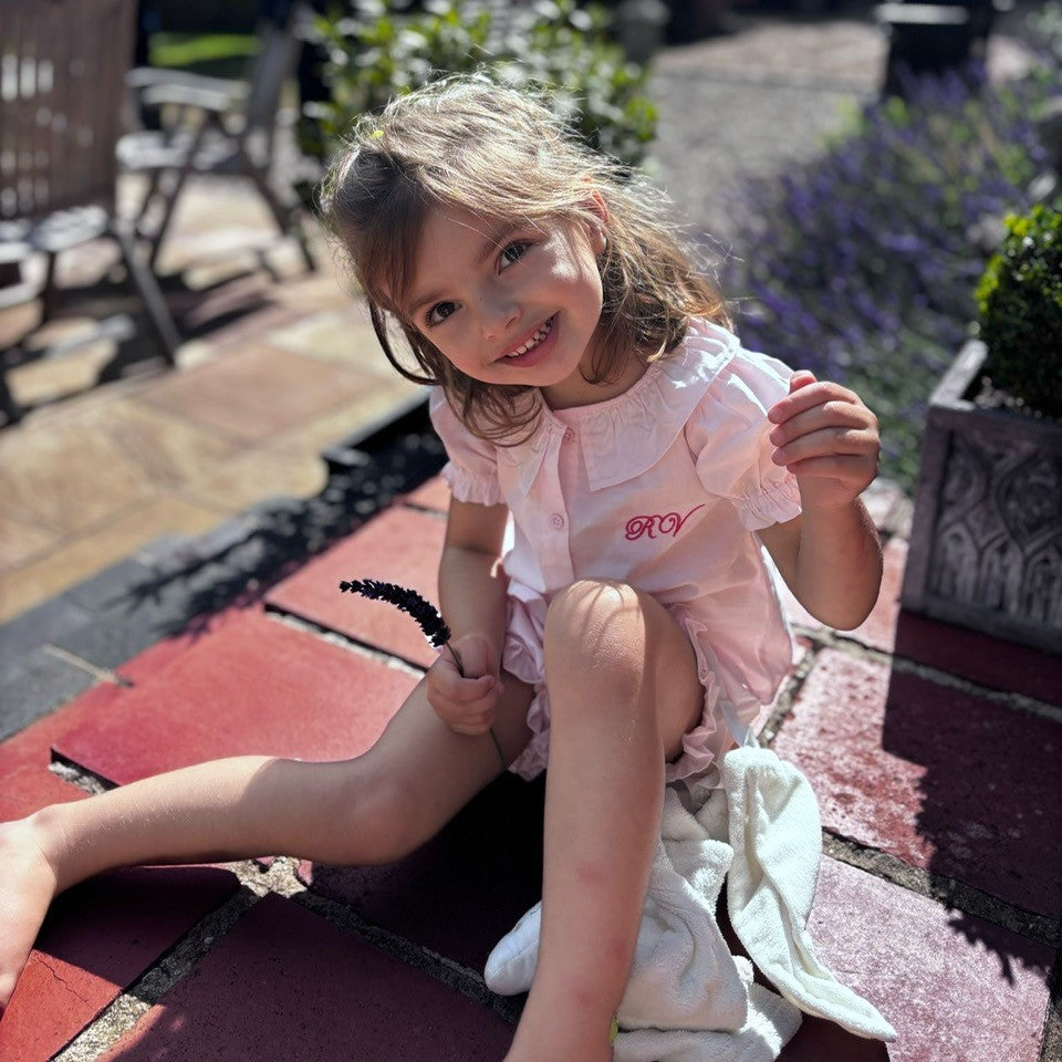 Young girl in a pink outift with personlised initials sitting on a red bench outdoors with plants in the background