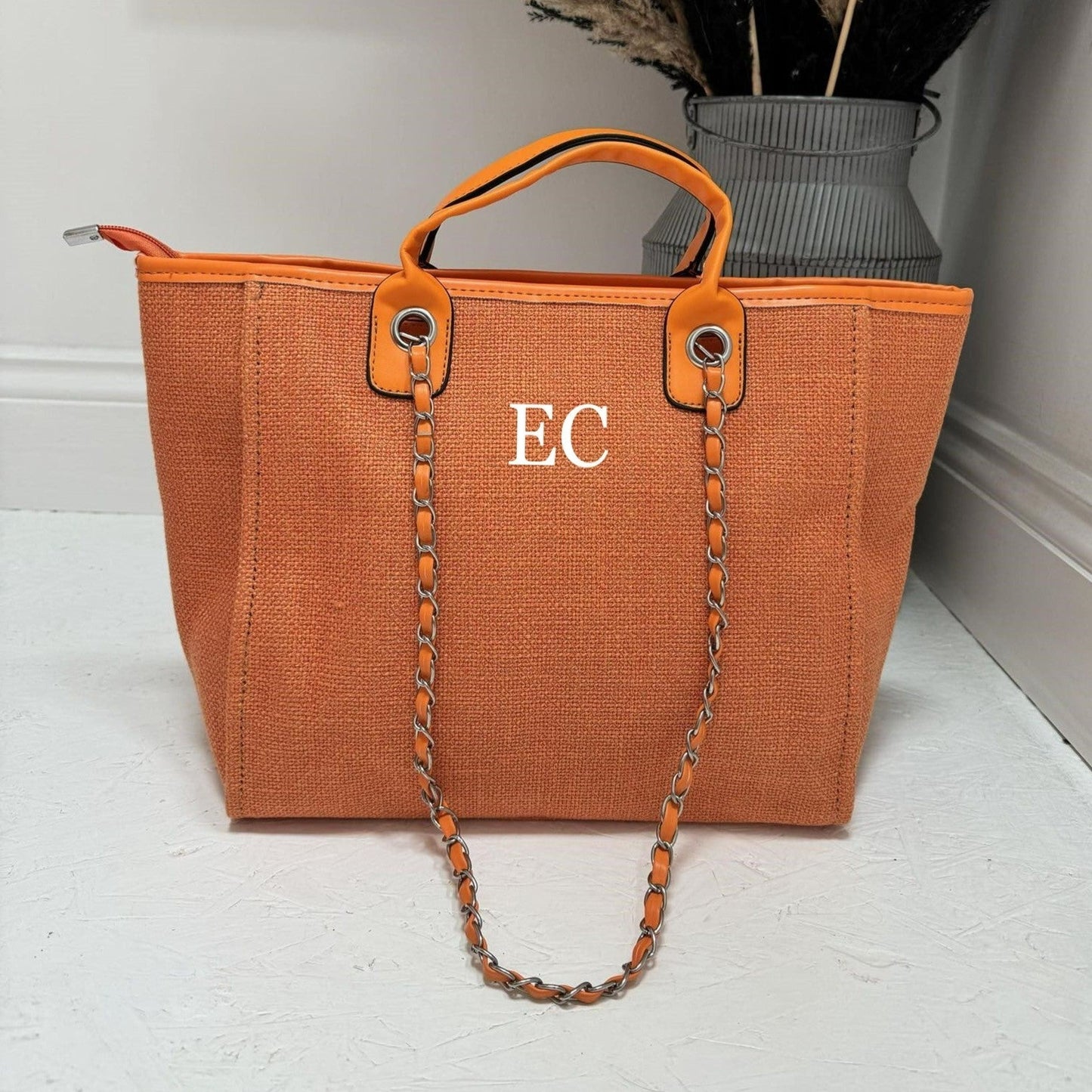 An orange tote bag with the initials 'EC' printed on the front, featuring a chain handle and a zipper closure.