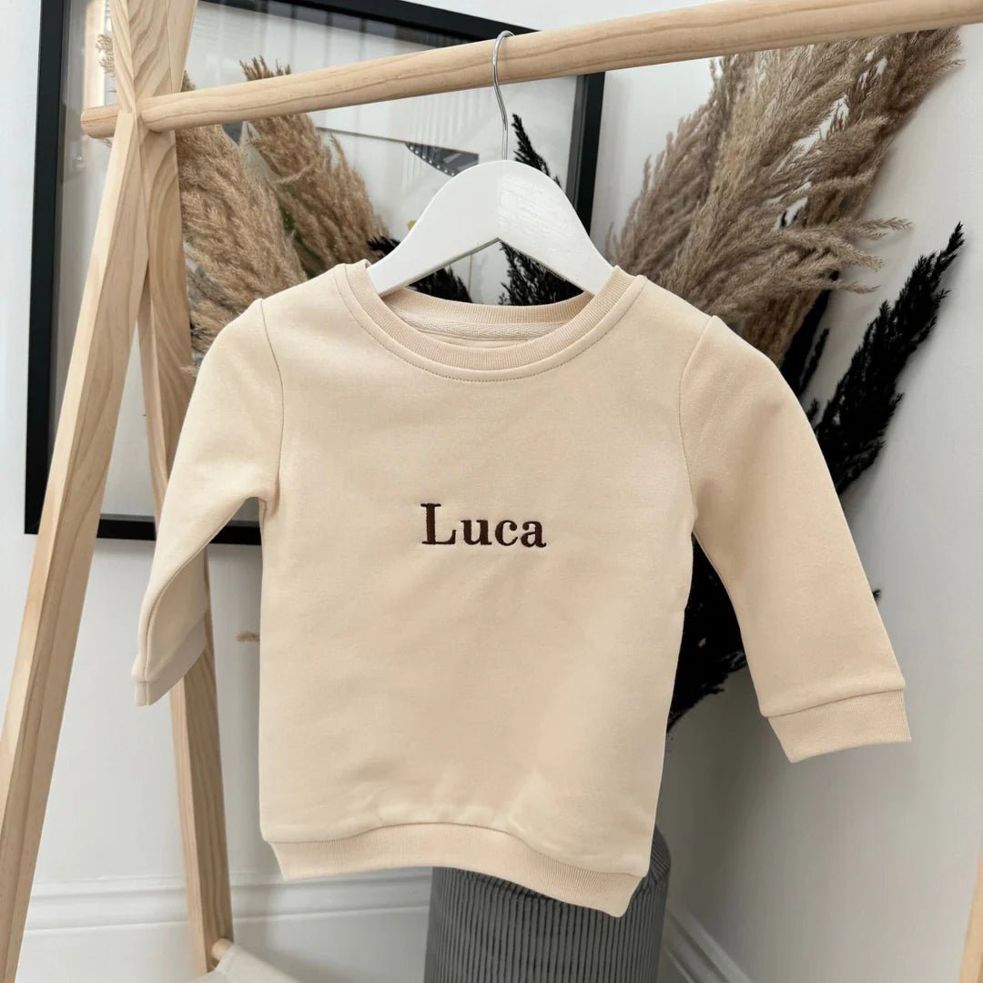 Beige sweatshirt with 'Luca' embroidered on it hanging on a wooden rack.
