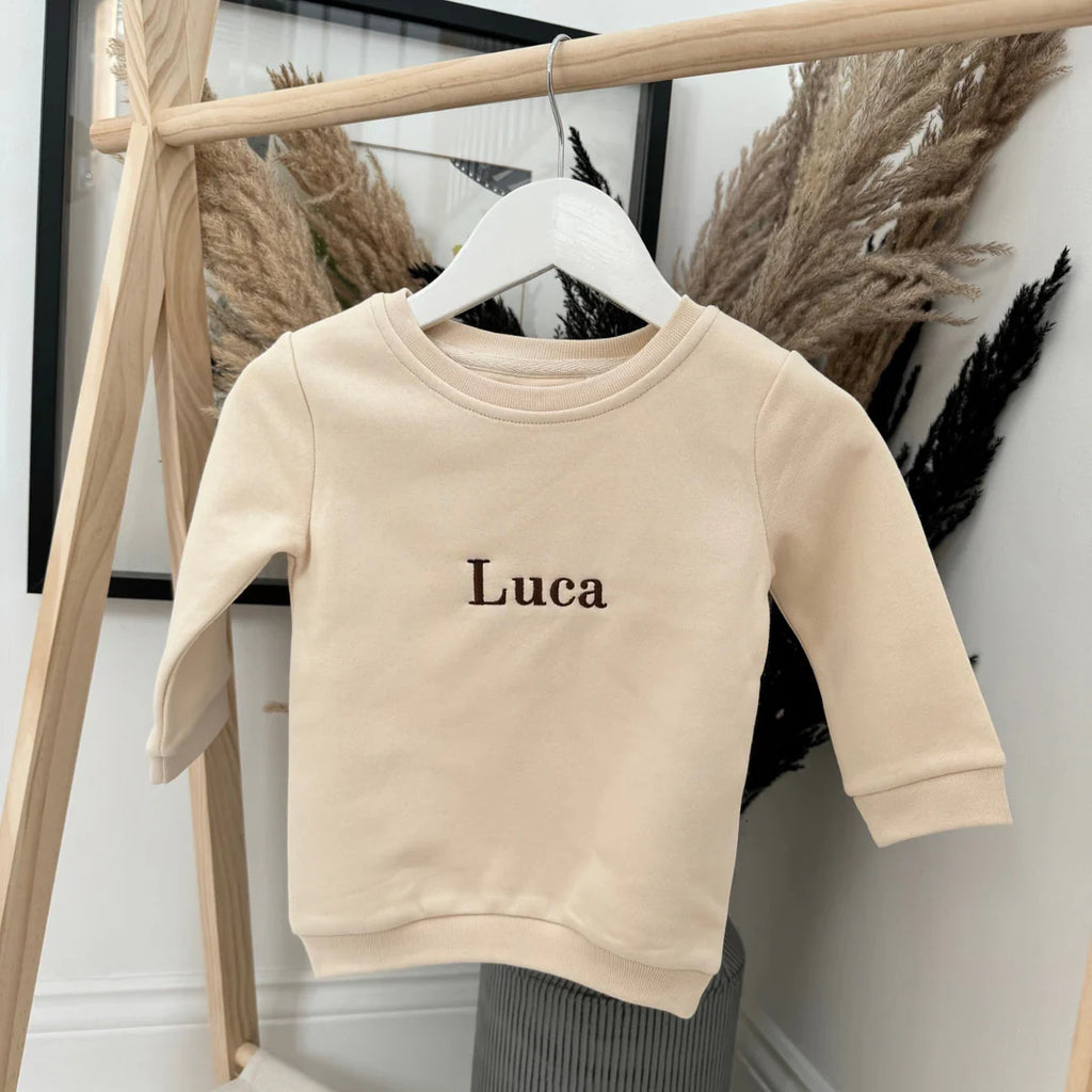 Beige sweatshirt with 'Luca' embroidered on it hanging on a wooden rack.