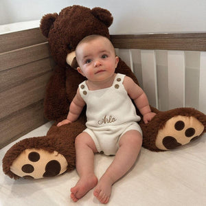 Baby in a crib with a large teddy bear, wearing a white romper with 'Arlo" embroidered.