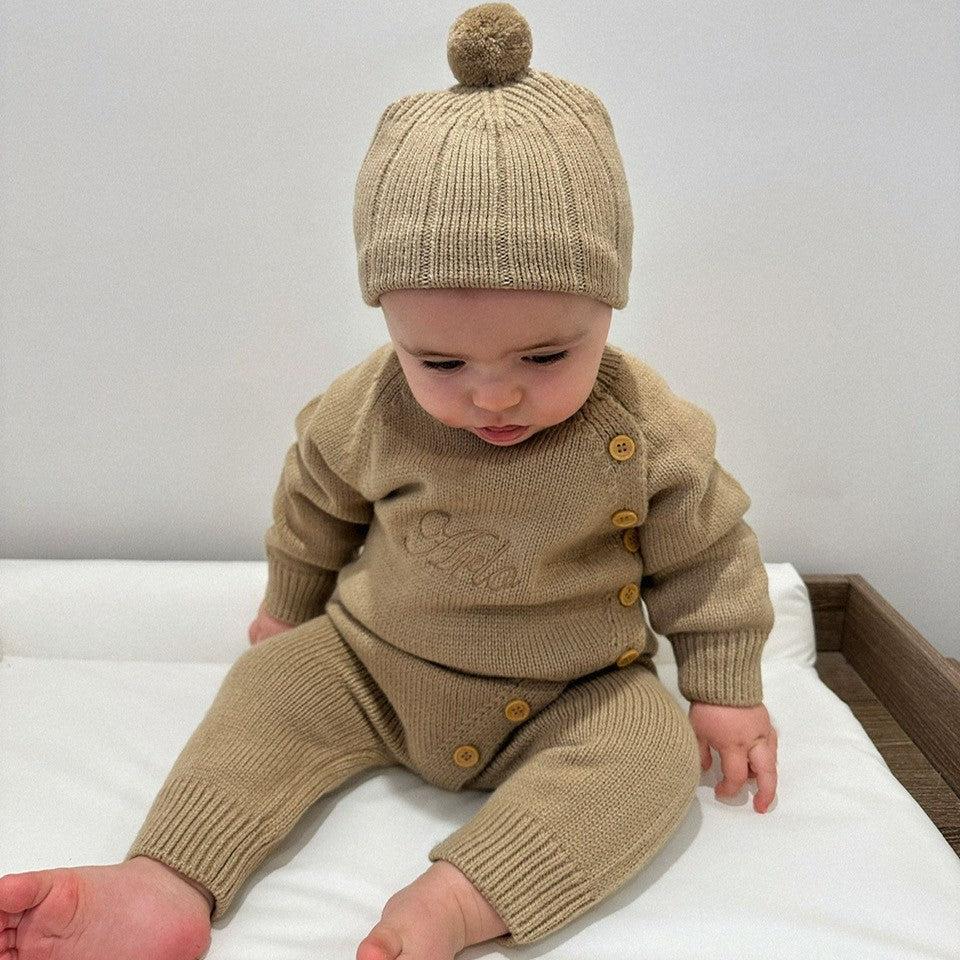 Baby wearing a brown knitted outfit with buttons and a matching hat on a white surface.