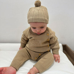 Baby wearing a brown knitted outfit with buttons and a matching hat on a white surface.