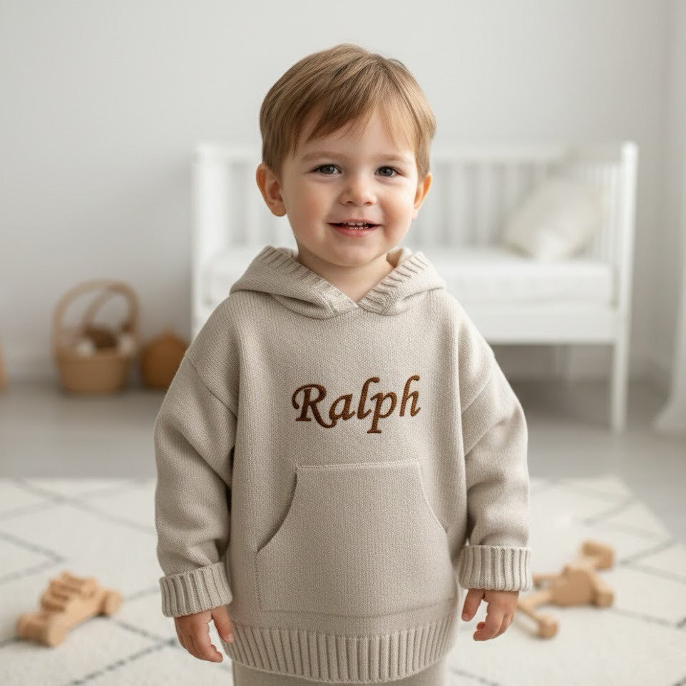 Child wearing a beige hoodie with 'Ralph' embroidered on it, standing in a bright room.