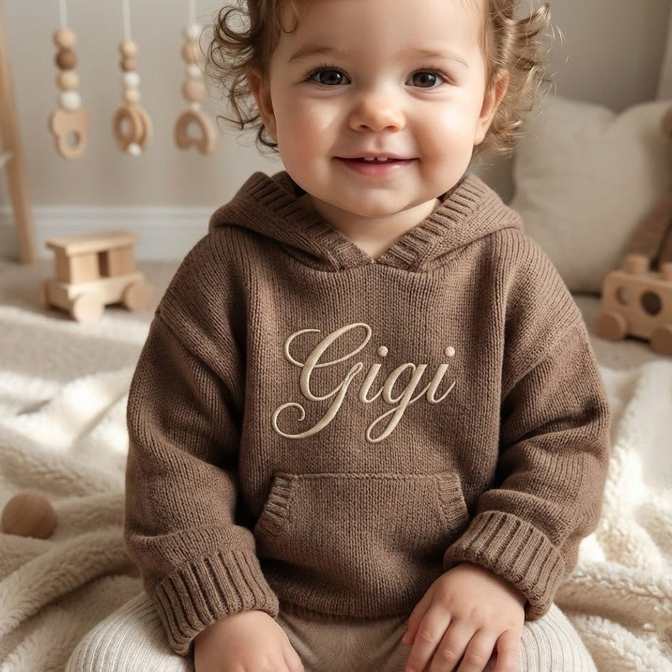 Child wearing a brown hoodie with 'Gigi' on it, sitting in a cozy room with toys.