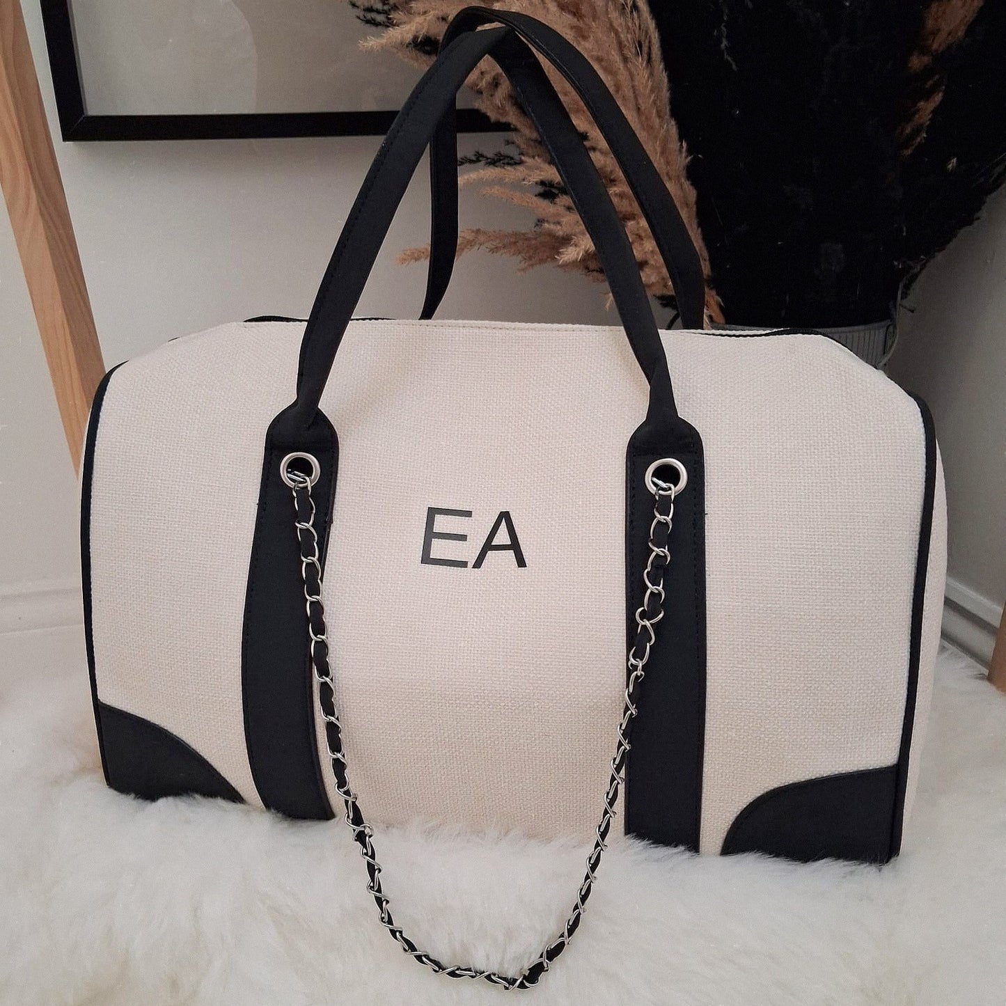 White duffel bag with black accents and a chain detail, featuring initials 'EA', on a light surface.