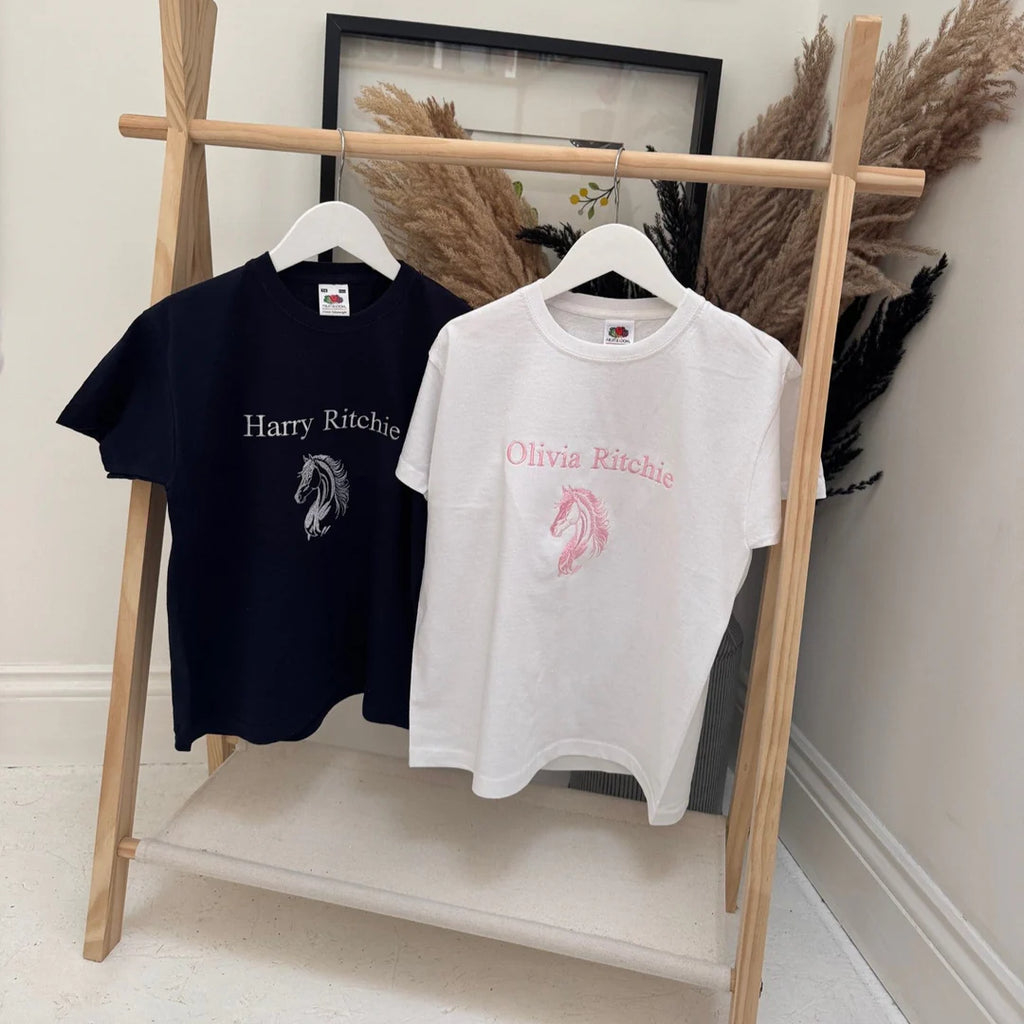 Two t-shirts, one navy and one white, with embroidered designs on a wooden rack.