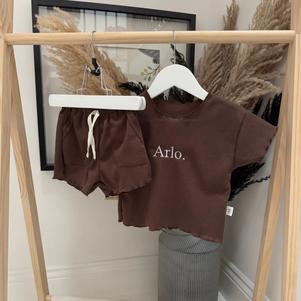 Brown children's outfit with 'Arlo' printed on a shirt hanging on a rack.