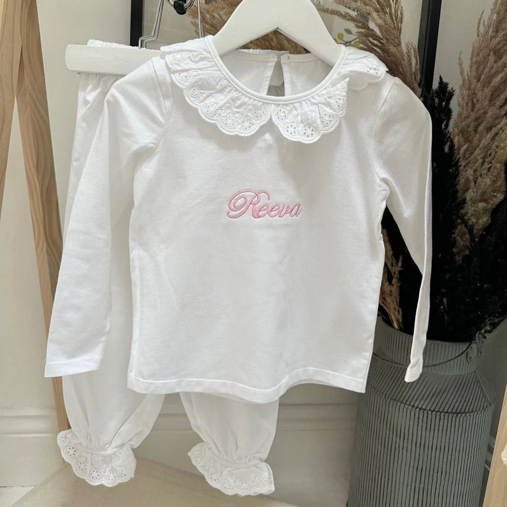 White baby outfit with lace details and 'Reeva' embroidery on a hanger.