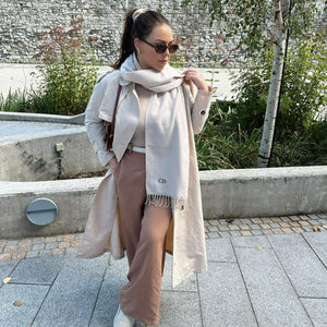 Woman wearing a long beige coat and scarf with sunglasses, standing outdoors.