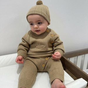 Baby wearing a beige knit outfit with a matching hat, sitting in a crib.