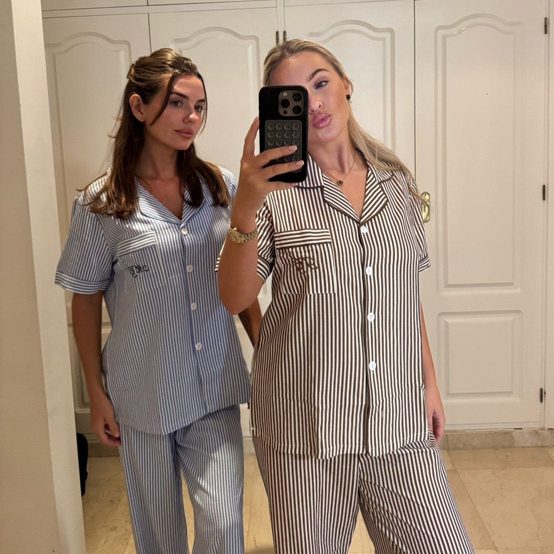 Two women taking a mirror selfie wearing matching striped pajama sets in a room with white cabinets.