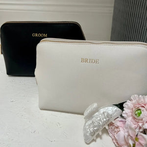 White pouch labeled 'BRIDE' with a black 'GROOM' pouch and flowers on a light surface.