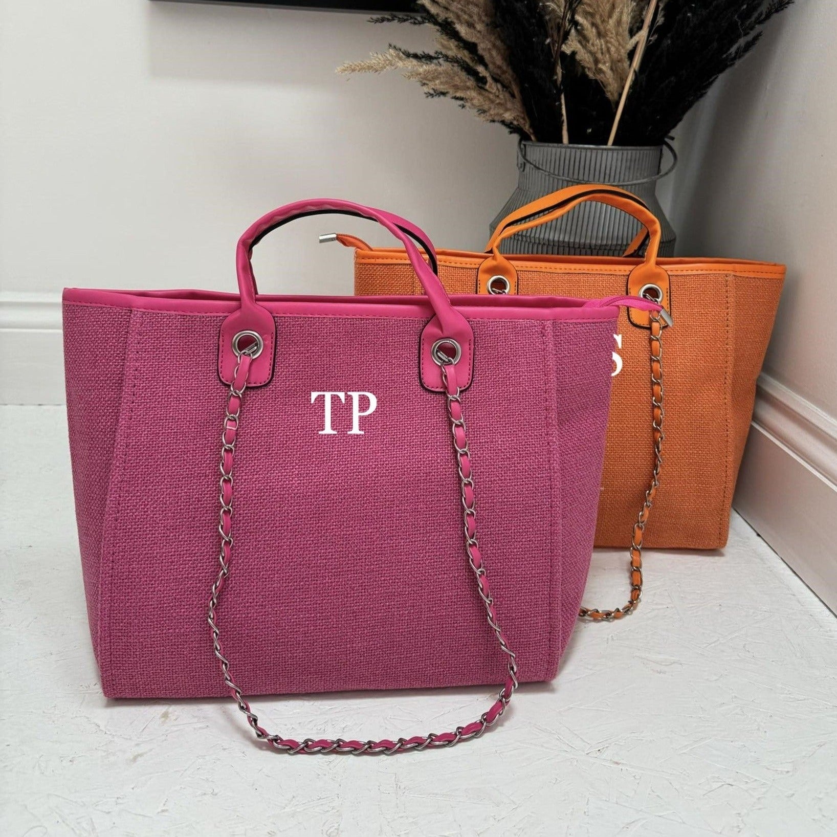 Two personalized tote bags one pink, one orange with shoulder straps, one in front of the other, on a bright surface. One bag has the initials 'TP' printed on the front.