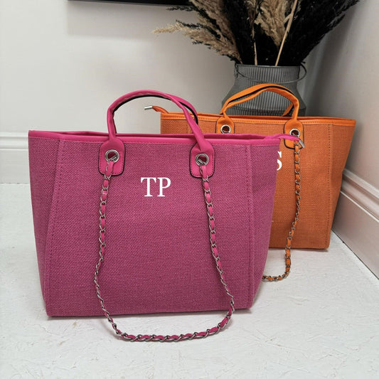 Two personalized tote bags one pink, one orange with shoulder straps, one in front of the other, on a bright surface. One bag has the initials 'TP' printed on the front.
