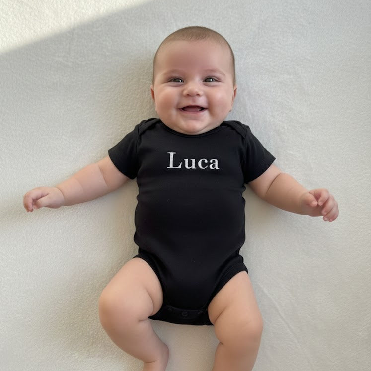 Baby wearing a black onesie with 'Luca' printed on it, lying on a light-colored surface.