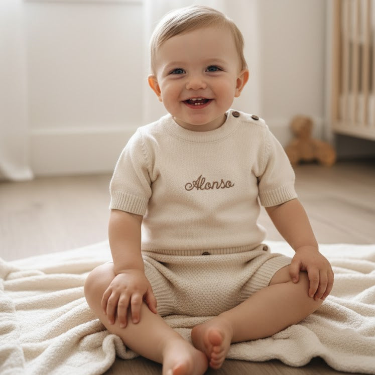 Baby sitting on a blanket in a room with a crib and toys.